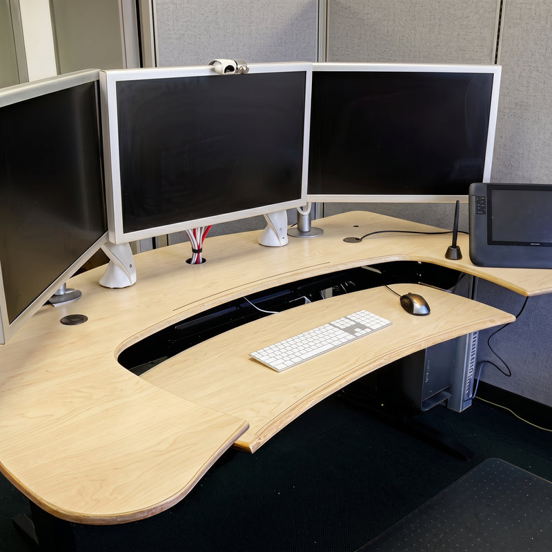 A modern curved standing desk with a light wood grain finish, featuring an integrated pull-out keyboard tray, a black under-desk tunnel, and a three-monitor monitor stand setup.