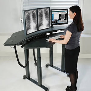 A woman stands at a curved, height-adjustable standing desk with dual computer monitors showing medical X-rays, in a bright clinical room.