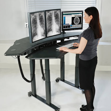 A woman stands at a curved, height-adjustable standing desk with dual computer monitors showing medical X-rays, in a bright clinical room.
